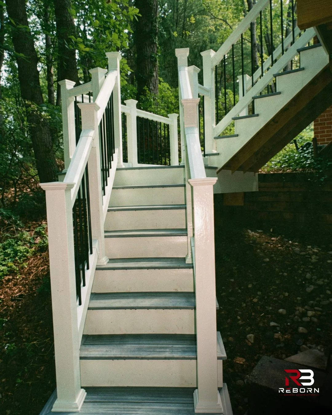 Outdoor deck stairs with white railings and black balusters, leading up into green trees. by Reborn General Construction LLC